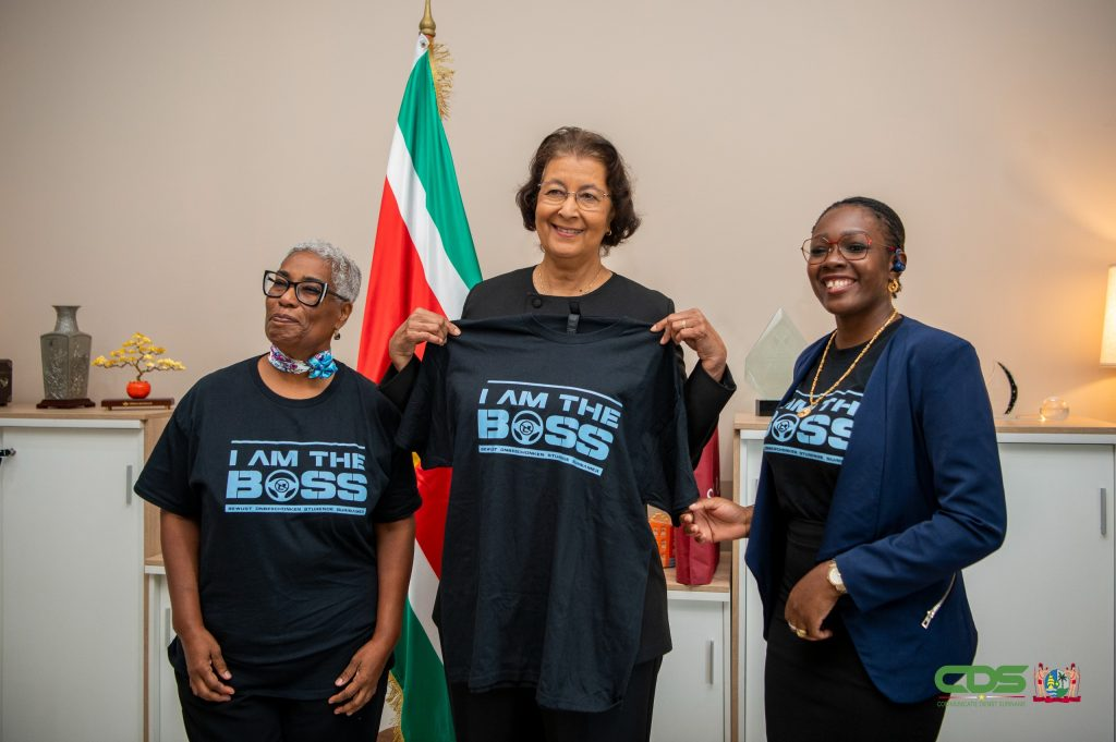 President Jennifer Simons houdt een BOSS-campagneshirt omhoog, met naast haar de directeur van het VVI en de voorzitter van STIVASUR